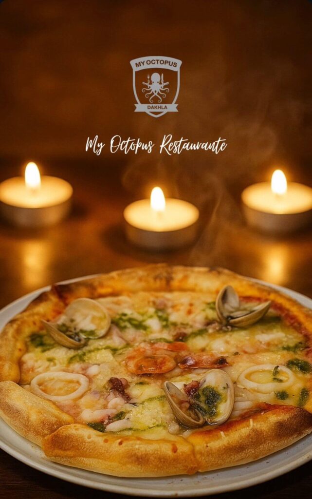 Candlelight Seafood Pizza My Octopus Dakhla A hot seafood pizza topped with clams, shrimp, and green sauce on a plate, with steam rising and three lit candles in the background, branded with My Octopus Dakhla logo.