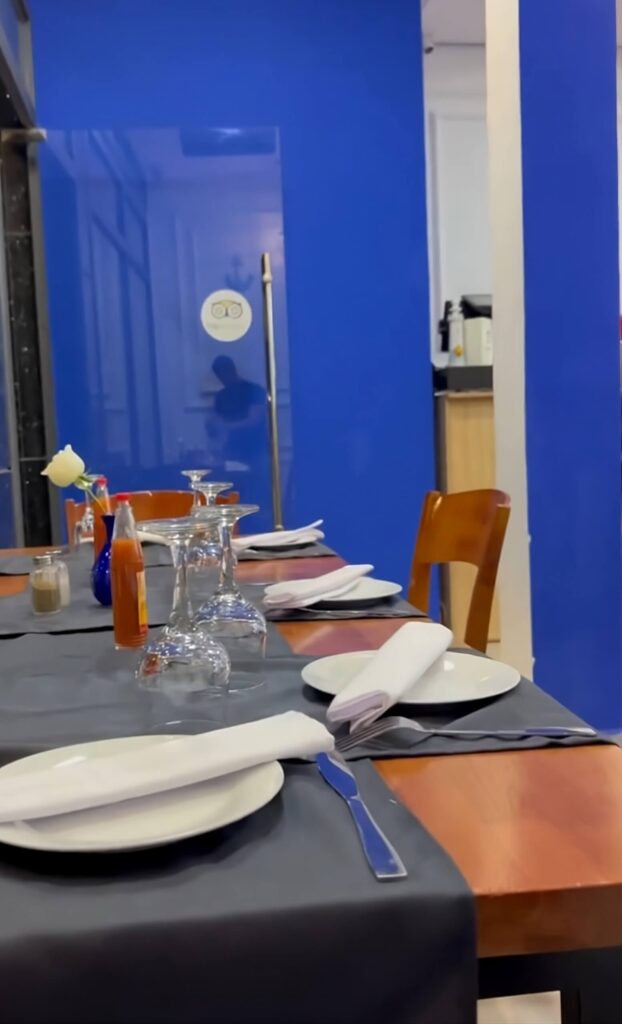 Ocean Blue Dining Experience My Octopus Dakhla A set dining table inside My Octopus Dakhla with white plates, folded napkins, upside-down glasses, and condiments, placed against bright blue walls and wooden chairs.